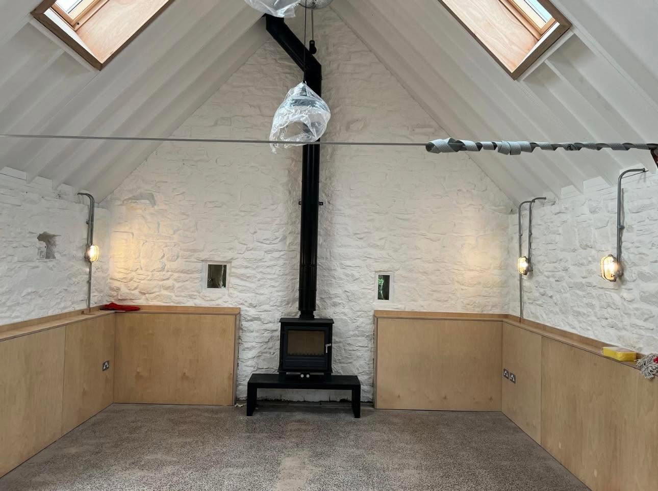 Interior with vaulted white painted stone walls, Velux rooflights, birch ply dado and wood burning stove