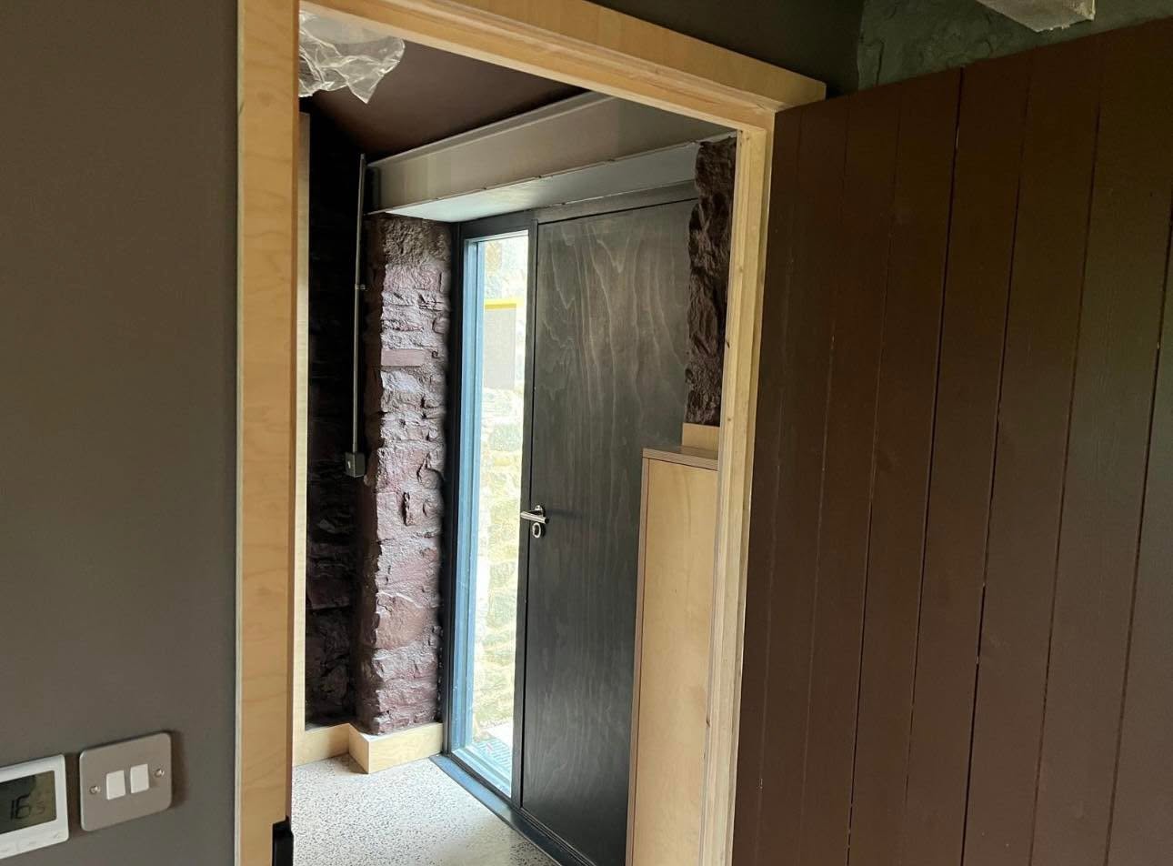 Entrance detail with ply lined opening through a thick stone wall to a black framed glazed door