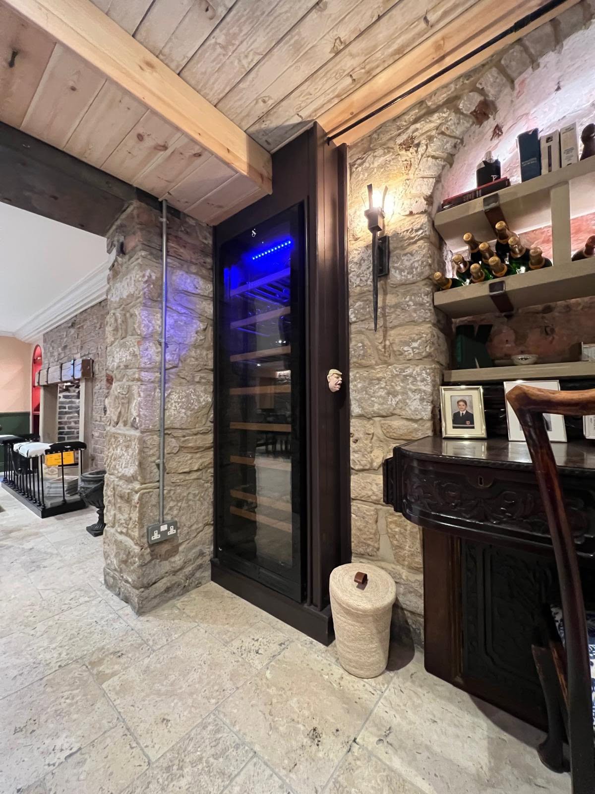 Integrated tall wine fridge framed in dark timber against stone pier with stone shelving and sconce