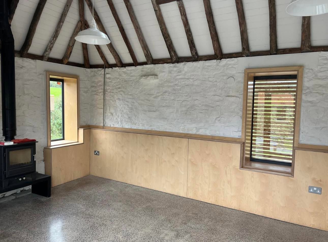 Interior with limewashed stone walls under retained rough timber rafters, birch ply dado and polished concrete floor