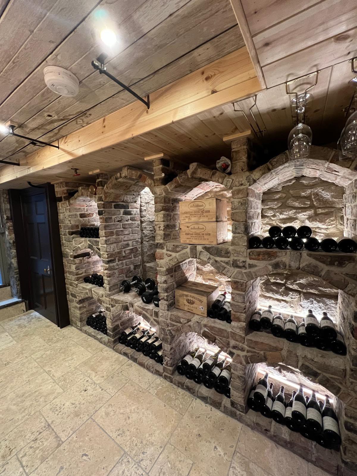 Wine cellar wide view with multiple arched bottle bays, lit shelves, timber ceiling and travertine floor
