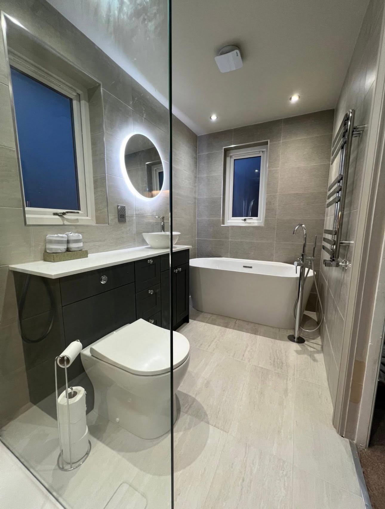 View past the shower screen showing freestanding bath, chrome towel radiator and dark shaker vanity run