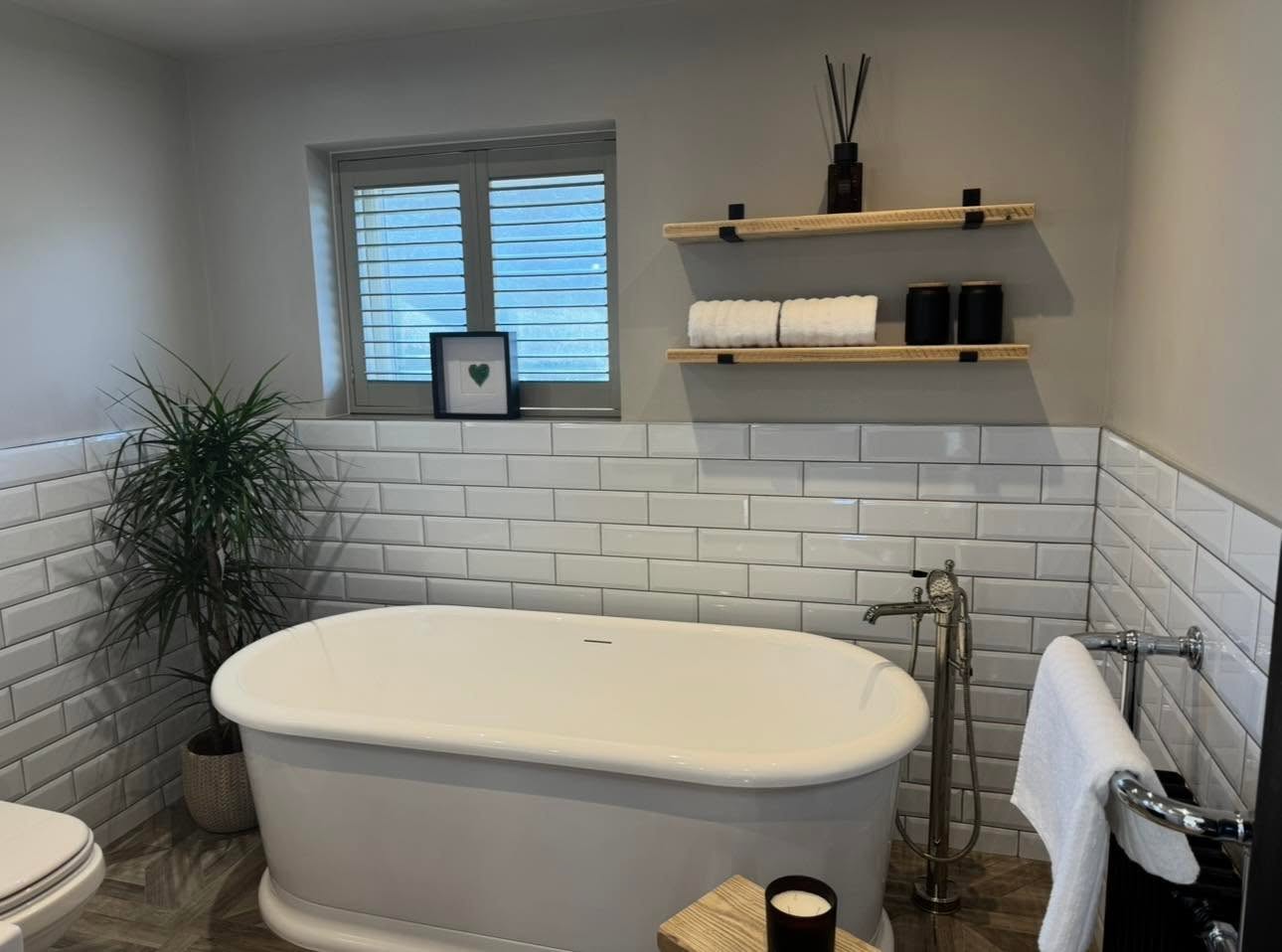 Oak floating shelves on black brackets beside the freestanding bath with metro tiling and shutters