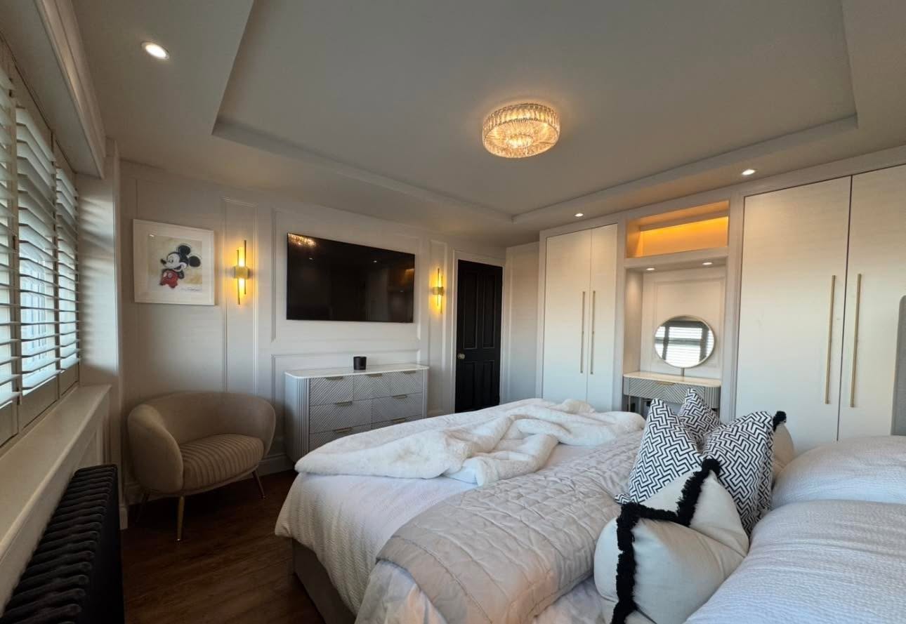 Master bedroom media wall with recessed TV in panelled surround, brass wall lights and fitted wardrobes with lit dressing nook