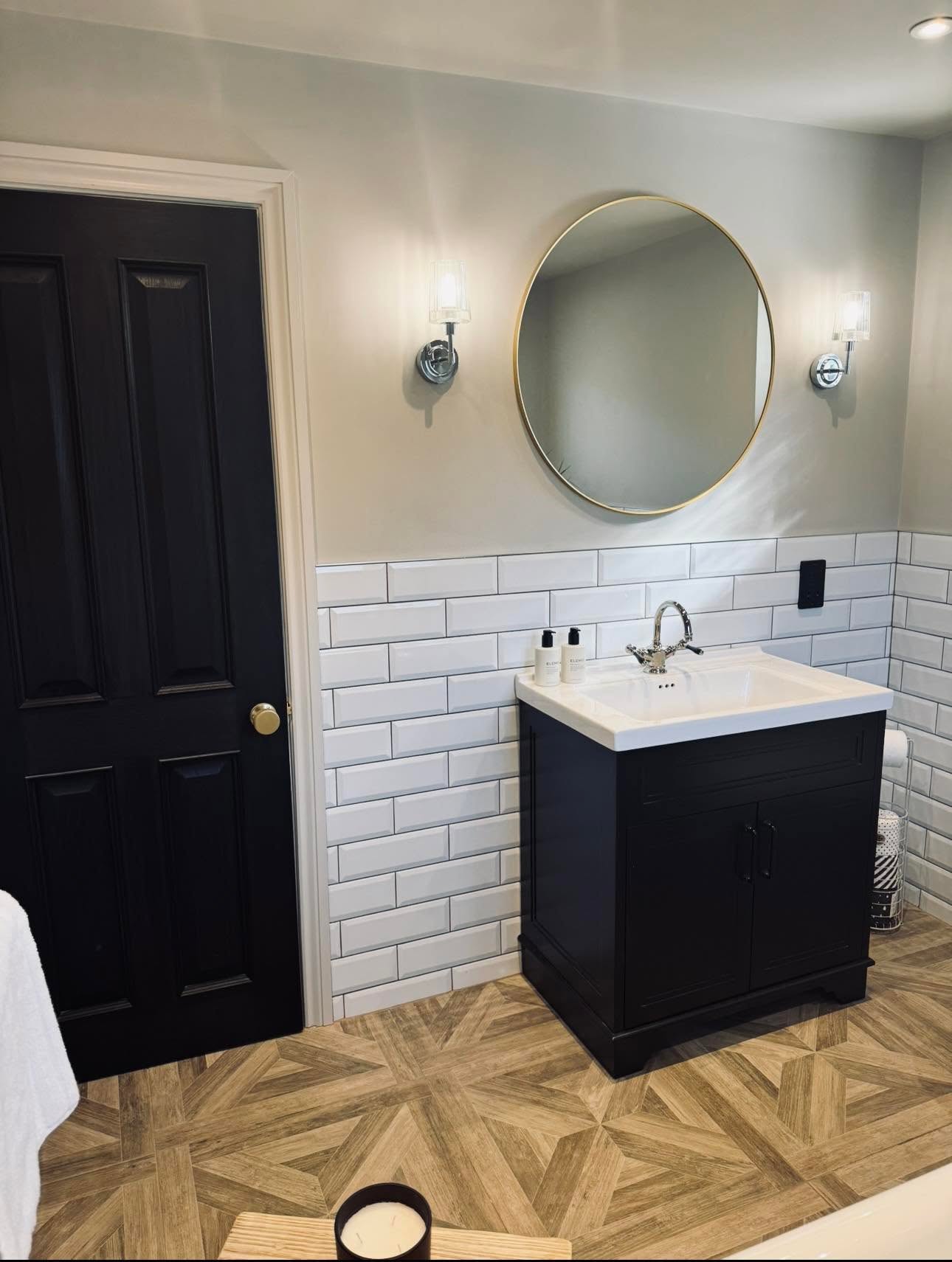Black painted vanity with white basin, round brass framed mirror and chrome wall lights against metro tiling
