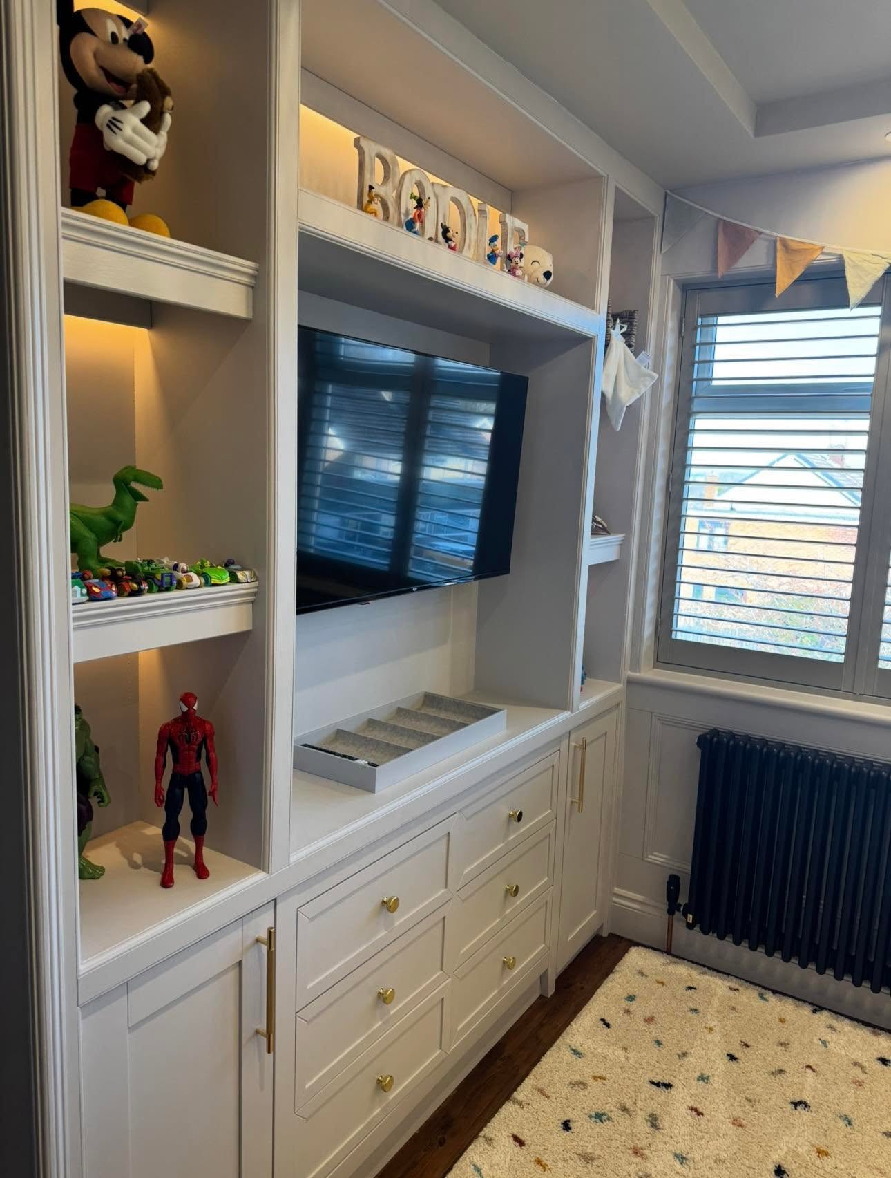 Child's bedroom media wall with fitted unit, recessed TV, lit display shelving, drawers with brass knobs and shutters